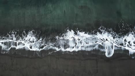 A-wave-hitting-the-black-sand-beach,-showcasing-the-contrast-of-white-foam-against-the-dark-shoreline,-aerial-view