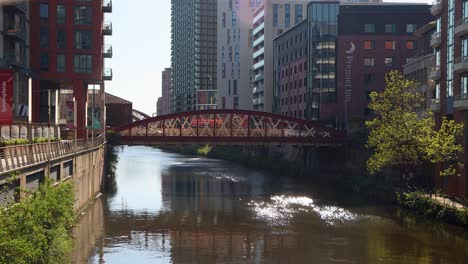 Sunny-day-view-of-red-bridge-over-a-calm-river-in-Manchester,-UK,-surrounded-by-modern-buildings