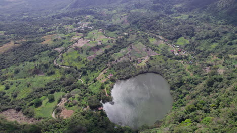 A-lush-green-landscape-with-a-circular-lake,-surrounded-by-dense-trees-and-patches-of-cultivated-land,-aerial-view