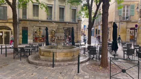 Quaint-European-cafe-scene-with-stone-fountain,-empty-chairs,-and-pedestrians-in-Aix-en-provence,-daylight