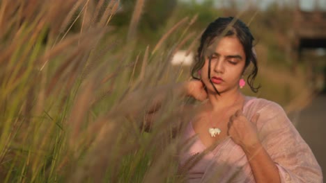 Young-woman-in-a-field-at-sunset,-touching-tall-grass,-with-warm-light-casting-a-glow-on-her-face