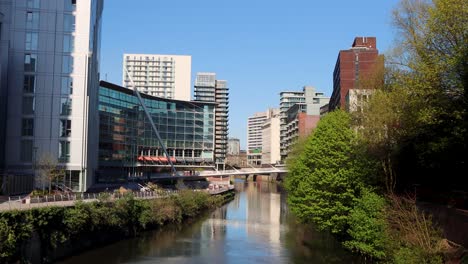 Sonniger-Tagesblick-Auf-Das-Stadtbild-Von-Manchester-Mit-Modernen-Gebäuden-An-Einem-Ruhigen-Fluss