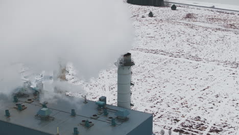 Industrial-smokestacks-emitting-steam-against-a-snowy-landscape,-overcast-weather,-aerial-view