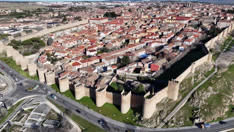 Rising-scramble-flight-viewing-the-medieval-walled-city-of-Avila-UNESCO-World-Heritage-Site-seeing-its-houses-inside-the-wall-and-a-road-with-cars-in-circulation-on-a-sunny-winter-day-in-Spain