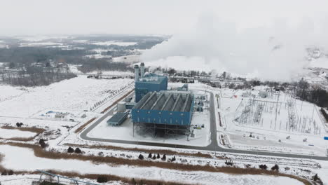 Eine-Industrieanlage-In-Einer-Verschneiten-Landschaft-Mit-Aufsteigendem-Dampf,-Winter,-Luftaufnahme