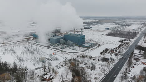 Industrial-power-plant-emitting-steam-in-a-snowy-landscape,-highway-in-foreground,-aerial-view