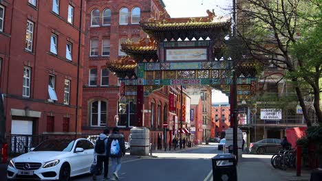 Peatones-Caminando-Bajo-La-Ornamentada-Puerta-De-Chinatown-En-Manchester-Durante-El-Día,-Cielo-Despejado