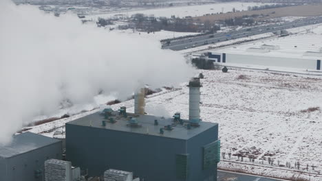 Industrial-buildings-with-smokestacks-in-winter,-emitting-steam-against-a-snowy-landscape,-aerial-view