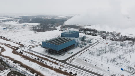 Industrial-plant-in-winter-landscape,-snow-covered-with-steam-rising-from-structures,-aerial-view