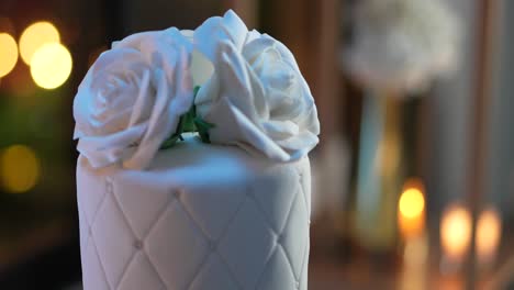 Wedding-cake-made-of-white-fondant,-with-edible-pearls-and-large-roses-on-top,-romantic-decorated-atmosphere-with-candles