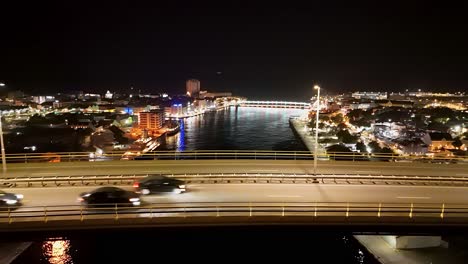Aerial-tracking-pan-across-Queen-Juliana-Bridge-as-cars-zoom-by-at-night,-Handelskade-WIllemstad-Curacao-in-background