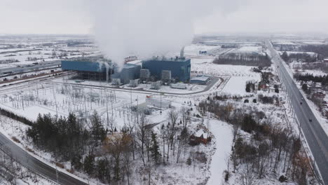 An-industrial-power-plant-in-a-snowy-landscape-with-active-smokestacks,-roads-and-trees,-aerial-view