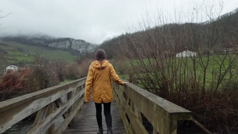 Excursionista-Con-Chaqueta-Amarilla-En-Un-Puente-Rural-Brumoso-Y-Caminando-Sobre-Una-Montaña-Brumosa