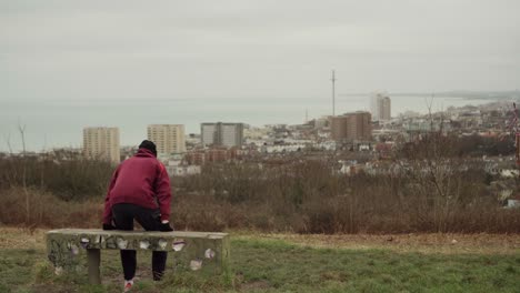 White-Man-in-Red-Hoodie-sits-on-bench-and-looks-over-Brighton,-Seaside-CIty