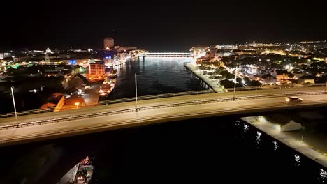 Die-Drohne-Senkt-Sich-Nachts-Unter-Die-Queen-Juliana-Bridge,-Während-Lichter-Auf-Dem-Wasser-über-Der-Handelskade-Curacao-Leuchten