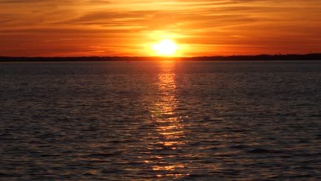 Glowing-sun-in-orange-sky-setting-beyond-calm-rippling-sea-horizon