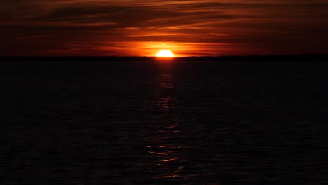 Sun-setting-beyond-dark-rippling-sea-horizon,-glowing-in-dim-sky
