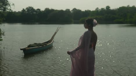 Woman-in-vintage-dress-by-river-with-canoe,-reflecting,-serene-mood,-daylight