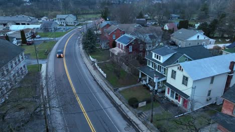 Drone-establishing-shot-of-white-car-driving-on-main-road-in-small-american-town-at-dusk