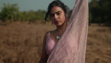 Woman-in-pink-sari-standing-in-dry-field,-intense-gaze,-sunlit-backdrop,-close-up