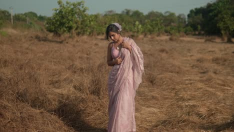 Woman-in-pink-saree-standing-thoughtfully-in-dry-grass-field,-under-clear-skies