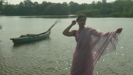 Woman-in-pink-saree-by-the-river,-adjusting-her-attire,-with-a-traditional-boat-in-the-background,-daylight