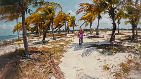A-female-riding-a-bike-along-a-beach,-under-palm-trees-on-a-sunny-day,-in-Caye-Caulker,-Belize