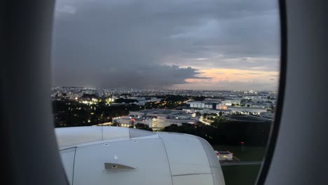 Vista-Aérea-A-Través-De-La-Ventana-Del-Avión-De-Los-Edificios-De-La-Ciudad-En-Singapur-Al-Atardecer-Con-Relámpagos-Y-Nubes