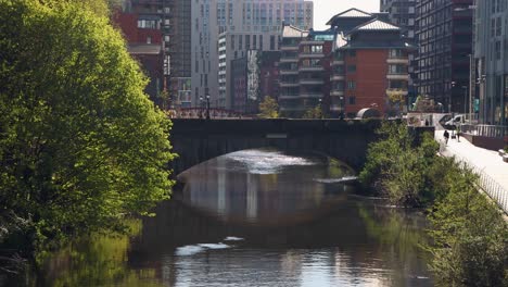 Sunny-view-of-bridge-over-river-in-Manchester-with-city-buildings-in-the-background-and-lush-greenery,-daytime