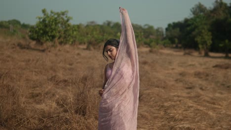 Woman-in-pink-sari-standing-in-dry-grass-field-with-cloth-over-head,-serene-mood,-daytime