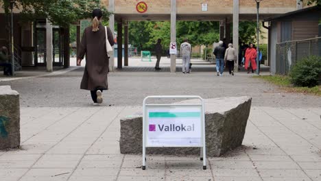 Pedestrians-walking-by-a-polling-station-sign-in-Stockholm,-election-atmosphere,-daytime,-urban-setting