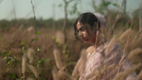 Woman-in-pink-dress-contemplating-in-dry-grass-field,-soft-focus,-warm-daylight