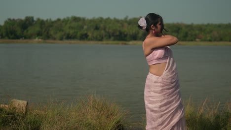 Woman-in-pink-saree-enjoying-breeze-by-river,-grass-in-foreground,-sunny-day