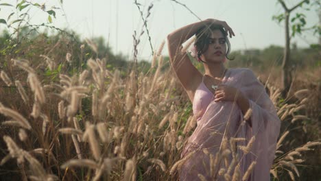 Frau-In-Rosa-Kleid-Posiert-Nachdenklich-In-Einem-Sonnenbeschienenen-Feld-Mit-Hohem-Gras,-Weicher-Fokus,-Natürliches-Licht