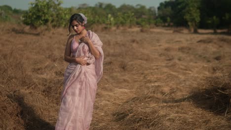 Woman-in-pink-saree-standing-thoughtfully-in-a-dry-grass-field,-trees-in-background,-daytime