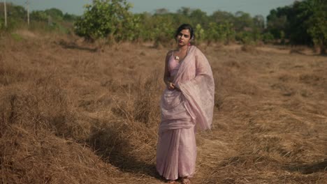 Woman-in-traditional-sari-standing-contemplatively-in-dry-grassland,-with-trees-in-the-background,-daylight