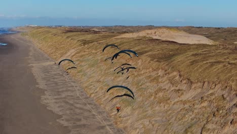 Group-of-paragliders-soar-along-grassy-sand-dune-slopes-of-Castricum-beach,-Netherlands