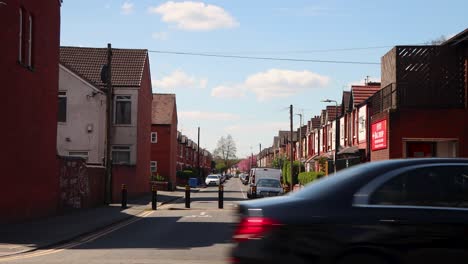 Row-of-red-brick-houses-under-blue-sky-in-Manchester,-pedestrian-crossing-the-street,-parked-cars,-sunny-day