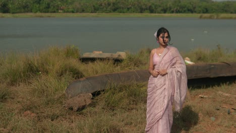 Woman-in-pink-saree-standing-by-a-river,-looking-over-shoulder,-with-lush-greenery-in-background