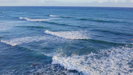 drone-footage-heading-off-shore-showing-the-white-capped-ocean-waves-of-the-Blue-Pacific-ocean-water-with-a-spattering-of-surfers-and-kite-boarders-awaiting-a-wave-near-Oahu-Hawaii