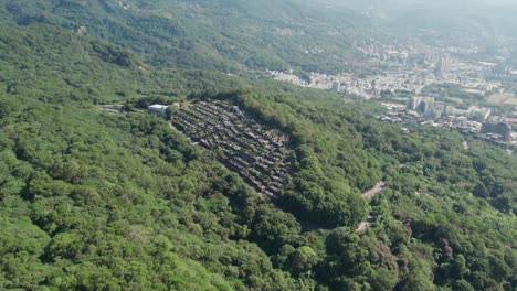 A-traditional-Taiwanese-cemetery-nestled-in-a-lush-green-forest-with-cityscape-in-the-background,-Aerial