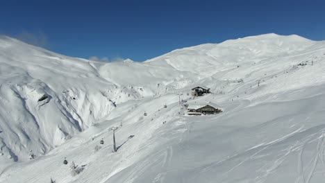 Vuelo-De-Drones-Sobre-Chalet-Y-Telesilla-Con-Esquiadores-En-La-Soleada-Ladera-De-La-Montaña-Bajo-Un-Cielo-Azul