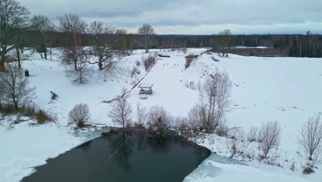 Aerial-drone-shot-over-a-pond-partially-frozen-surrounded-by-snow-covered-landscape-on-a-cold-winter-day