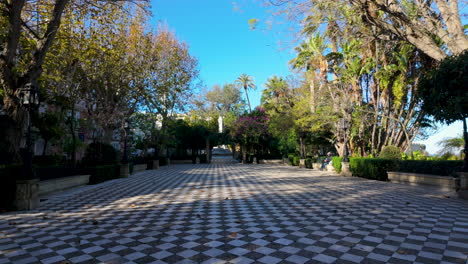 A-wide-pathway-lined-with-diverse-trees-and-palms-in-a-tranquil-public-park,-with-clear-blue-skies,-inviting-for-a-leisurely-walk-or-a-peaceful-retreat-in-an-urban-environment