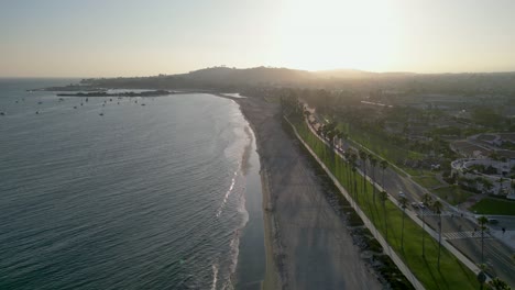 Drone-flying-up-Santa-Barbara,-CA-coastline-at-sunset-with-footage-of-ocean-waves-crashing-gently-against-the-beach-and-people-walking-on-the-shoreline