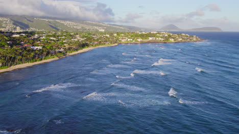 Las-Olas-Cubiertas-De-Blanco-Encabezan-El-Agua-Azul-Del-Océano-Del-Pacífico-A-Lo-Largo-De-La-Costa-De-La-Isla-De-Oahu,-Hawaii,-Con-Pueblos-Costeros-Y-Crecimiento-Tropical.