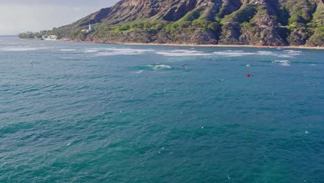 as-the-drone-backs-away-from-the-shore-of-Diamond-head-on-the-island-of-Oahu-surfers-and-kite-boarders-enjoy-gliding-on-the-turquoise-waters-surface-in-Hawaii