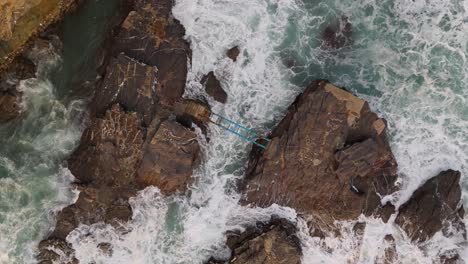 Waves-crashing-on-rocks-with-a-blue-footbridge-in-nervi,-genoa,-italy,-aerial-view