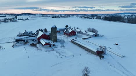 Sunrise-over-snowy-farm-with-red-barns,-silo,-and-bare-trees