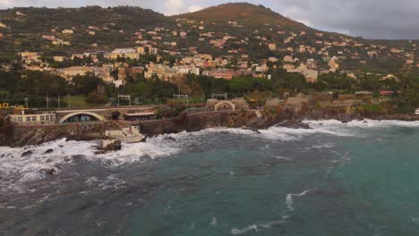 Rugged-coastline-and-waves-crashing-against-rocks-with-genoa-city-in-background,-overcast-sky,-aerial-view